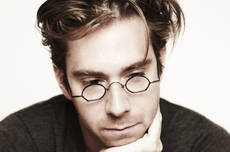 A headshot of a man with brown hair and circular glasses. He is gazing downwards with his chin resting in his hand.