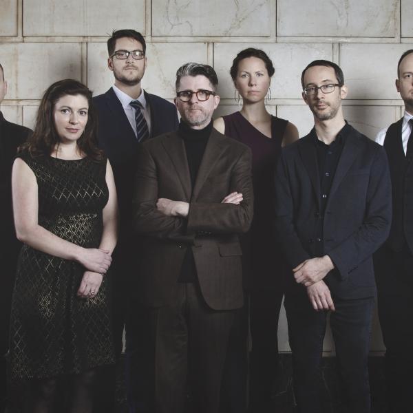 Wet Ink ensemble – Five men and two women, wearing black, stand in front of a block wall