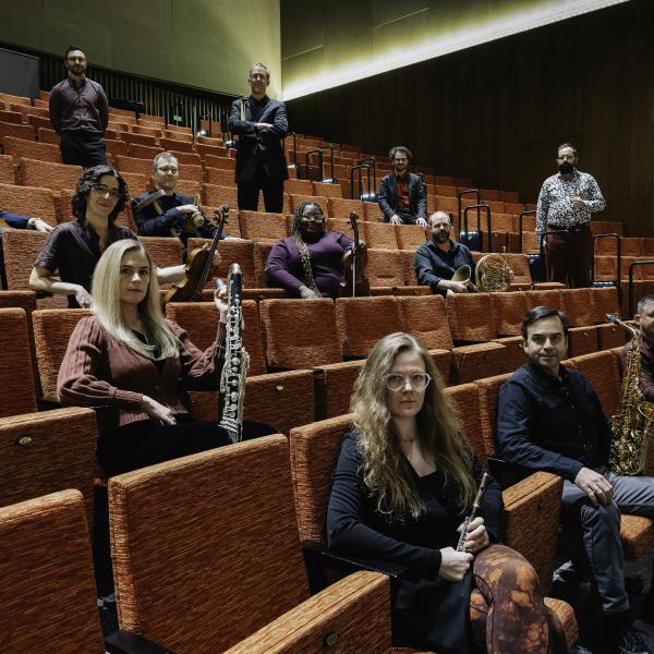 A group of musicians sits in the seats of a performance hall. They are sitting spaced out in different rows and seats, staring powerfully at the camera with their instruments.